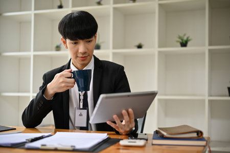 A smart and handsome young Asian male office worker or banker in a formal business suit is sipping coffee while reading something on his tablet at his desk in the office.の写真素材
