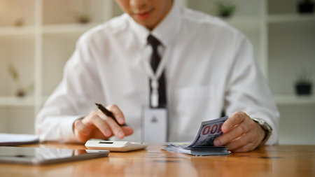 Close-up image of a professional Asian male banker using a calculator and counting US dollar bills at his desk in the office. financial investment, savings, currency exchangeの写真素材