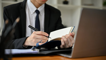 Cropped image of a professional Asian male lawyer in a formal suit writing something on his notebook while working on his lawsuit at his desk in the office.の写真素材