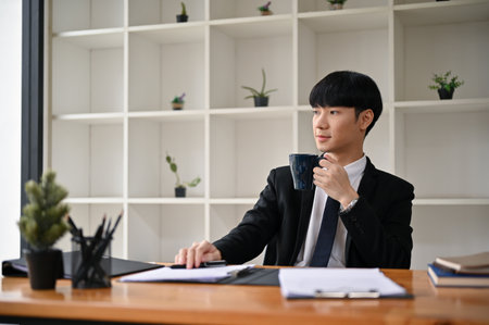A handsome and successful millennial Asian male lawyer or legal consultant in a formal suit is sipping his morning coffee at his desk.の写真素材