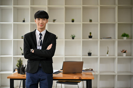 A handsome and smart young Asian businessman or male office worker in a business suit stands in his office with his arms crossed and looks at the camera.の写真素材