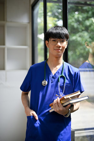 A portrait of a smart and professional young Asian male doctor in scrubs and glasses stands in the hospital corridor with books.の写真素材