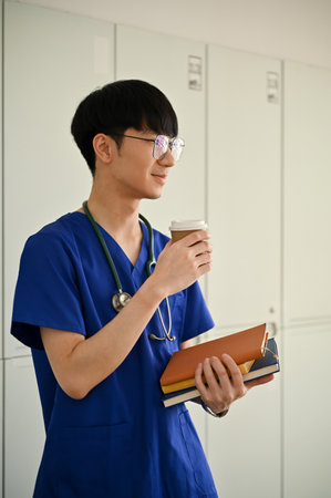 A portrait of a handsome young Asian male medical student or doctor in scrubs stands in the hospital corridor with a coffee cup and books in his hands. career, job, health care conceptの写真素材