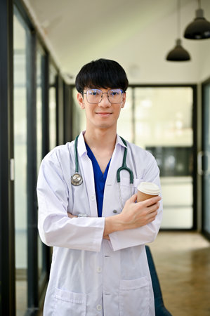 A portrait of a handsome and positive millennial Asian male doctor in a uniform with a stethoscope stands in the hospital corridor with a coffee cup in his hand.の写真素材