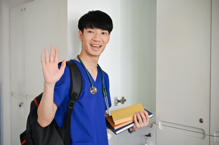 A positive and handsome young Asian male medical student in scrubs waving his hand, greeting or saying hi to his friends while walking in the locker room at the hospital.の写真素材