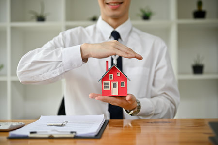Close-up image of a professional male real estate agent or realtor holding a house model at his desk. property investment, residential purchaseの写真素材