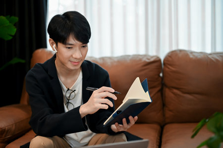 Relaxing young Asian man reading books and taking note while sitting on the sofa. Productive time for himselfの写真素材