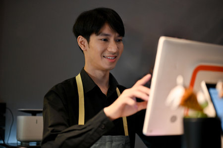 Young Asian male coffee shop owner checking bills for customer with machine. coffee shop worker concept, small businessの写真素材