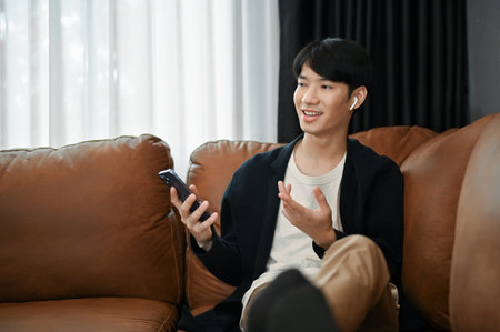 Relaxing young Asian man sitting oh their sofa and holding smartphone in his hand in free time.の写真素材