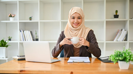 A portrait of a beautiful and successful Asian Muslim businesswoman or female boss in hijab, smiling and looking at the camera while sitting at her desk with a coffee cup in her hands.の写真素材