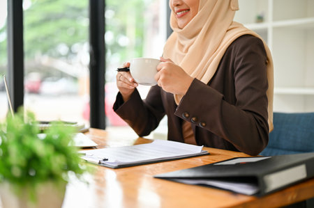 Close-up image of a successful and attractive Asian Muslim businesswoman in hijab sipping her morning coffee at her desk in the office.の写真素材
