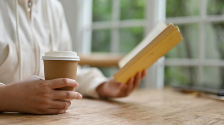 Close-up image of a woman in casual clothes sipping coffee and reading a book while relaxing in a coffee shop.の写真素材