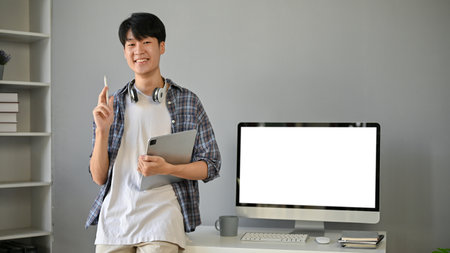 A handsome and happy young Asian man in a flannel shirt stands in his modern office with a tablet in his hand. a computer white screen mockup on a tableの写真素材