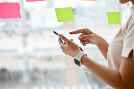 Close-up image of a woman using her smartphone while standing by the window in her office. text,...