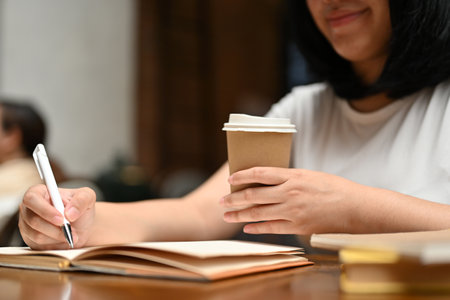 Close-up image of a happy young Asian woman sipping coffee and writing a diary while relaxing in a...
