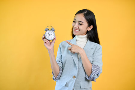 An attractive Asian businesswoman pointing at an alarm clock on her hand while standing against an isolated yellow background.の写真素材