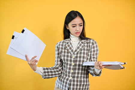 A confused and perplexed young Asian businesswoman looking nat documents on her hand while standing against an isolated yellow background.の写真素材