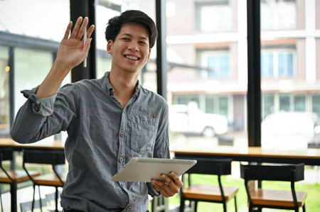 A handsome and happy young Asian man in casual clothes stands in a coffee shop with his portable digital tablet.の写真素材