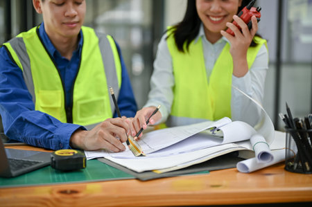Two professional Asian construction engineers wearing safety vests are checking a blueprint, discussing work, and working in the office together.の写真素材
