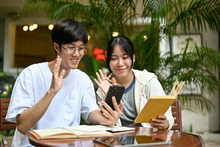 A happy young Asian male and female friend are enjoying talking on a video call with their friend while relaxing at a cafe together.の写真素材