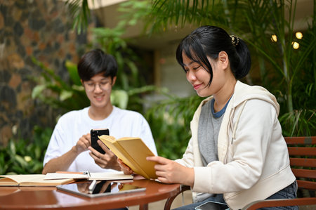 A pretty and happy young Asian female college student is reading a book while her male friend is using a smartphone, sitting at a cafe in an outdoor area together.の写真素材