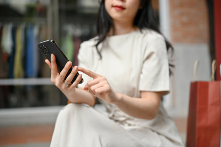 Close-up image of a pretty Asian woman texting with her friends on her smartphone while resting on the stairs in front of a clothing store after her shopping day in the city.の写真素材