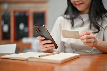 Close-up image of a beautiful young Asian woman using her smartphone and her credit card to pay...