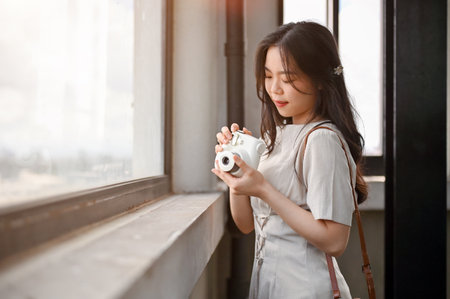 A beautiful Asian female tourist in casual clothes is standing by the window in a tower and taking pictures of cityscapes with her instant camera. Lifestyle conceptの写真素材