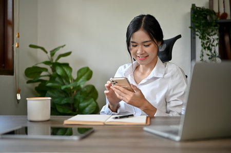 A beautiful Asian businesswoman texting with her friends or checking emails on her smartphone while sitting at her desk in her office.の写真素材