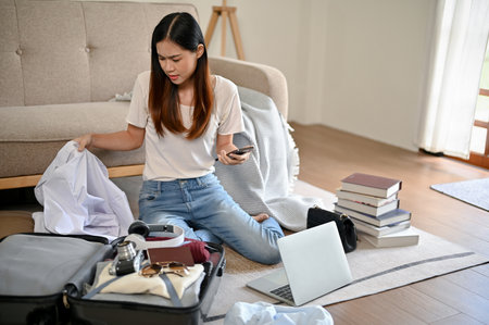 A busy young Asian woman is preparing her belongings and packing her luggage in her living room, getting ready for her trip abroad.の写真素材