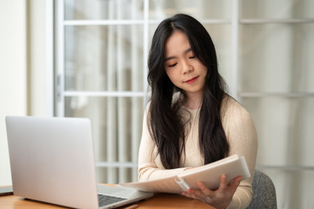 A beautiful young Asian female college student focuses on reading information in a book while studying online at home. Education and people conceptの写真素材