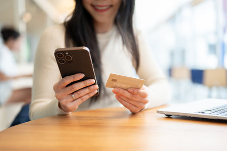 Close-up image of a happy and beautiful Asian woman using her mobile banking app while sitting at a table in a coffee shop. digital payment, cashless lifestyle, online banking, debit cardの写真素材