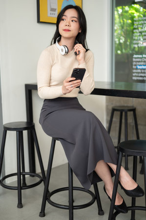 A portrait of a beautiful Asian woman in casual clothes with a smartphone in her hand is sitting at...