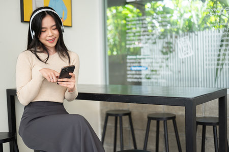 A beautiful and happy young Asian woman is listening to music on her headphones and using her smartphone while relaxing in a coffee shop.の写真素材