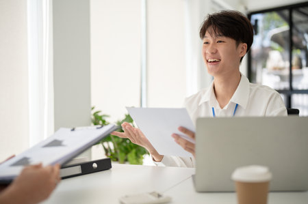 A cheerful and handsome young Asian male office worker discusses work and shares his ideas with a female colleague while working in the office together.の写真素材