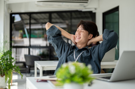 A happy and smiling young Asian businessman is leaning back in his chair, putting his hands behind his head, eyes closed, daydreaming, and feeling satisfied and carefree at work.の写真素材