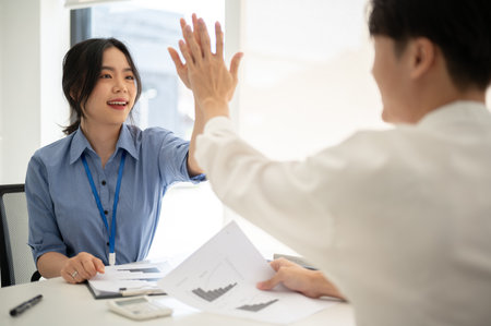 A cheerful young Asian businesswoman or female office worker gives high fives to a male colleague while working on a project together. teamwork, co-working, cheer-up, team buildingの写真素材
