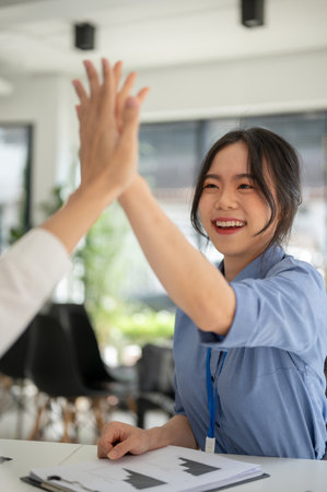 A portrait of a cheerful young Asian businesswoman gives high fives to a male colleague while...