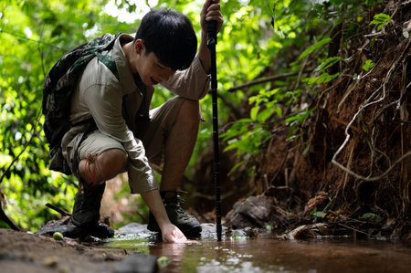 A happy Asian male hiker sits by the nature stream, enjoying nature, touching water with his hands,...