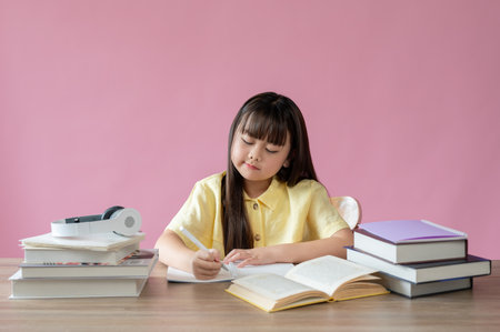 An adorable young Asian girl is focusing on her homework at her study table against an isolated pink background. Elementary school students conceptの写真素材