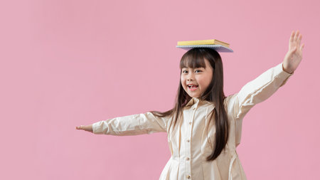 A cheerful and adorable young Asian girl puts books on her head and makes a flying gesture against an isolated pink studio background. freedom, imagination, carefree, happy kid, educationの写真素材