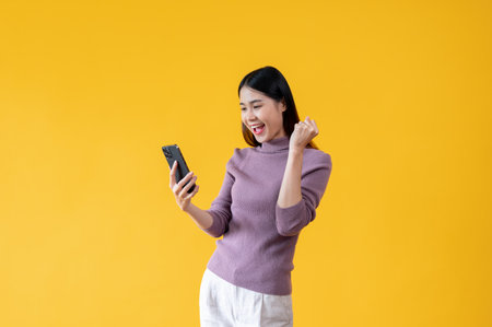 A cheerful young Asian woman is showing clenched fist and looking at her smartphone with a happy face while standing against an isolated yellow background. good news, hot promotion, special discountの写真素材
