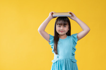 A cheerful and adorable young Asian girl is holding books over her head, smiling, and looking at the camera. isolated yellow background. Kindergarten, kids, students, elementary school studentsの写真素材