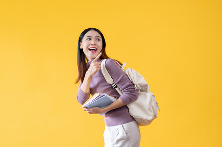 A cheerful and smiling young Asian female college student with her backpack is holding books and pointing her finger backward at an empty space on an isolated yellow background.の写真素材