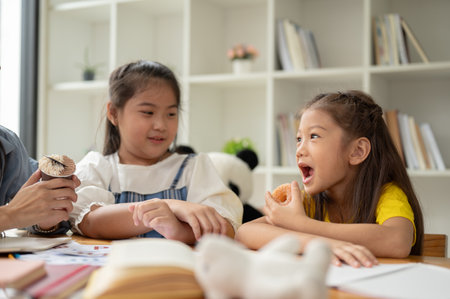 An adorable young Asian girl is eating a doughnut on her snack break while studying with her private teacher. children, kids, preschool, kindergarten studentsの写真素材