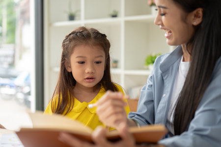 A pretty Asian preschool girl is studying and reading texts in a book with a kind and caring female teacher. Learning, studying, child development, parentingの写真素材