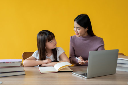 An adorable young Asian girl is studying with a female tutor, sitting at a table together. isolated yellow background. private tutor, home school, education conceptの写真素材