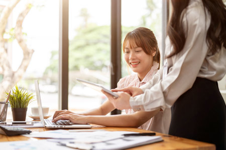 A pretty Asian businesswoman or female office worker is discussing work with her coworker while working on her tasks on a laptop at her desk in the office.の写真素材