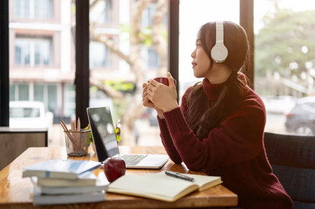 An attractive young Asian woman is listening to music on her headphones, looking out the window, and...