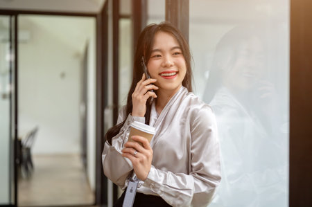 A portrait of an attractive and smiling Asian businesswoman is talking on the phone while standing in an office corridor with a coffee cup in her hand.の写真素材